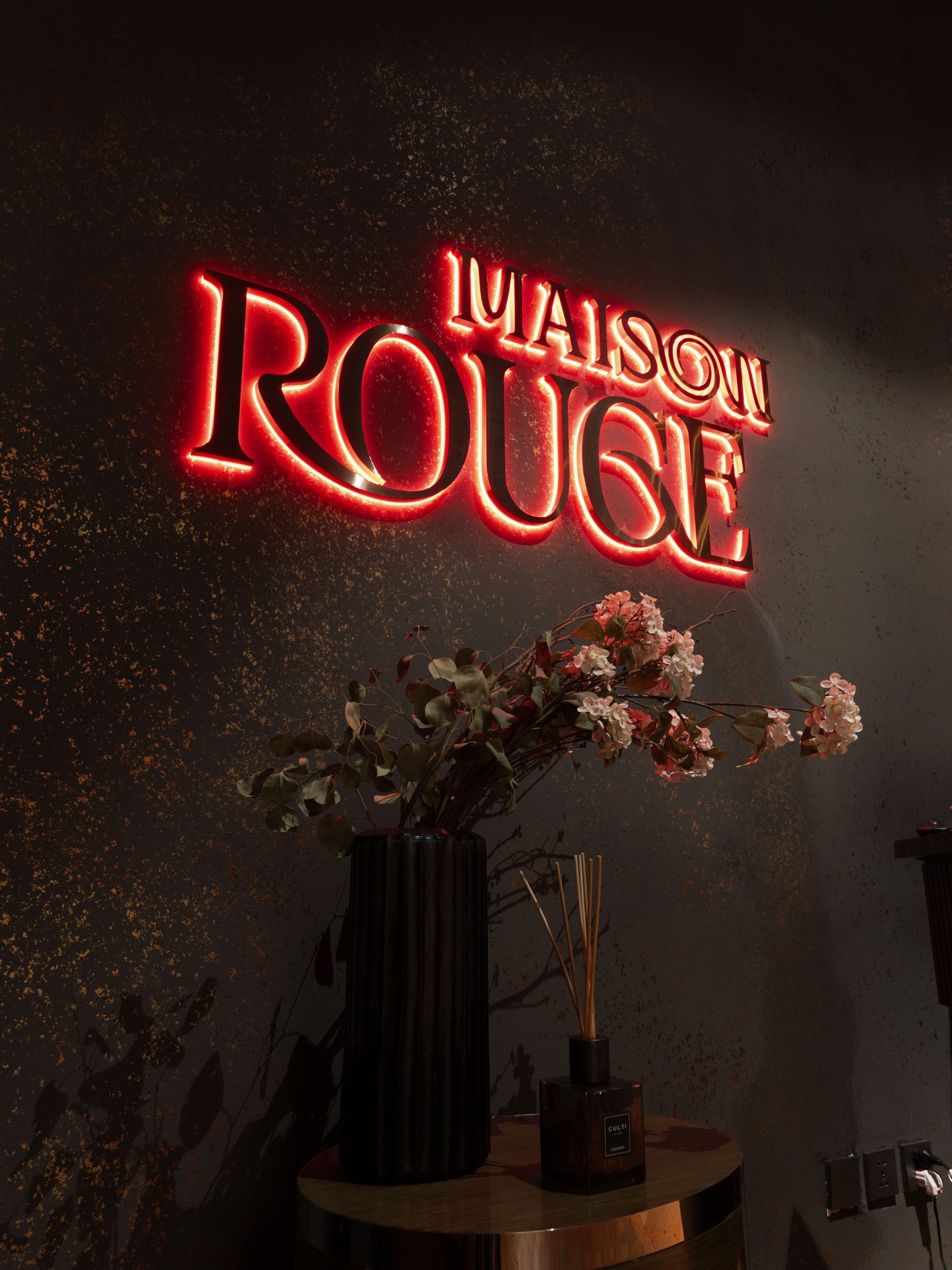 A red neon Maison Rouge sign above a slim console and cherry blossom stems